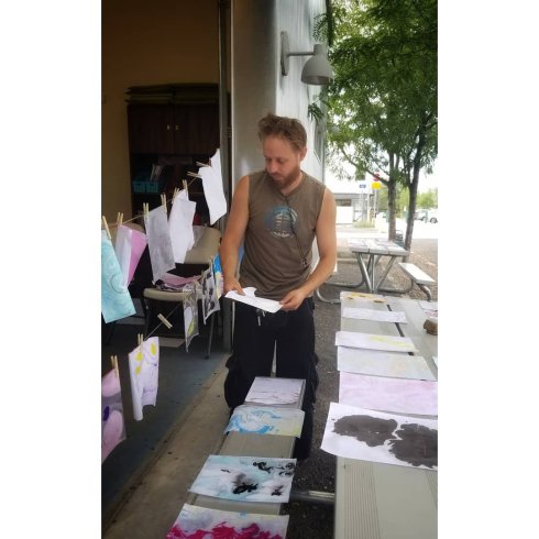 A white man with a beard and sleeveless shirt moves the drying paper from the bench to the clothesline